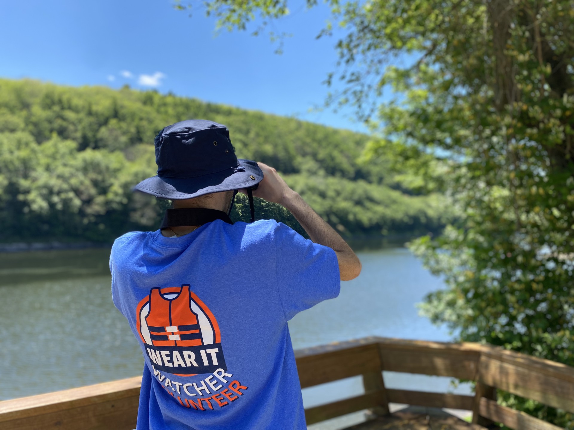 Yung man wearing blue "Wear It Watcher Volunteer" T-shirt looks at river with binoculars