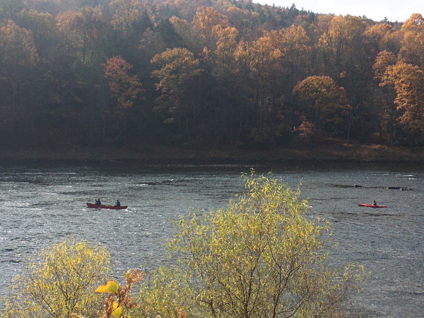 Trees in the background are red, yellow, and orange in color. A river runs right to left with two boaters on the water. Trees in the background are red, yellow, and orange in color. A river runs right to left with two boaters on the water.