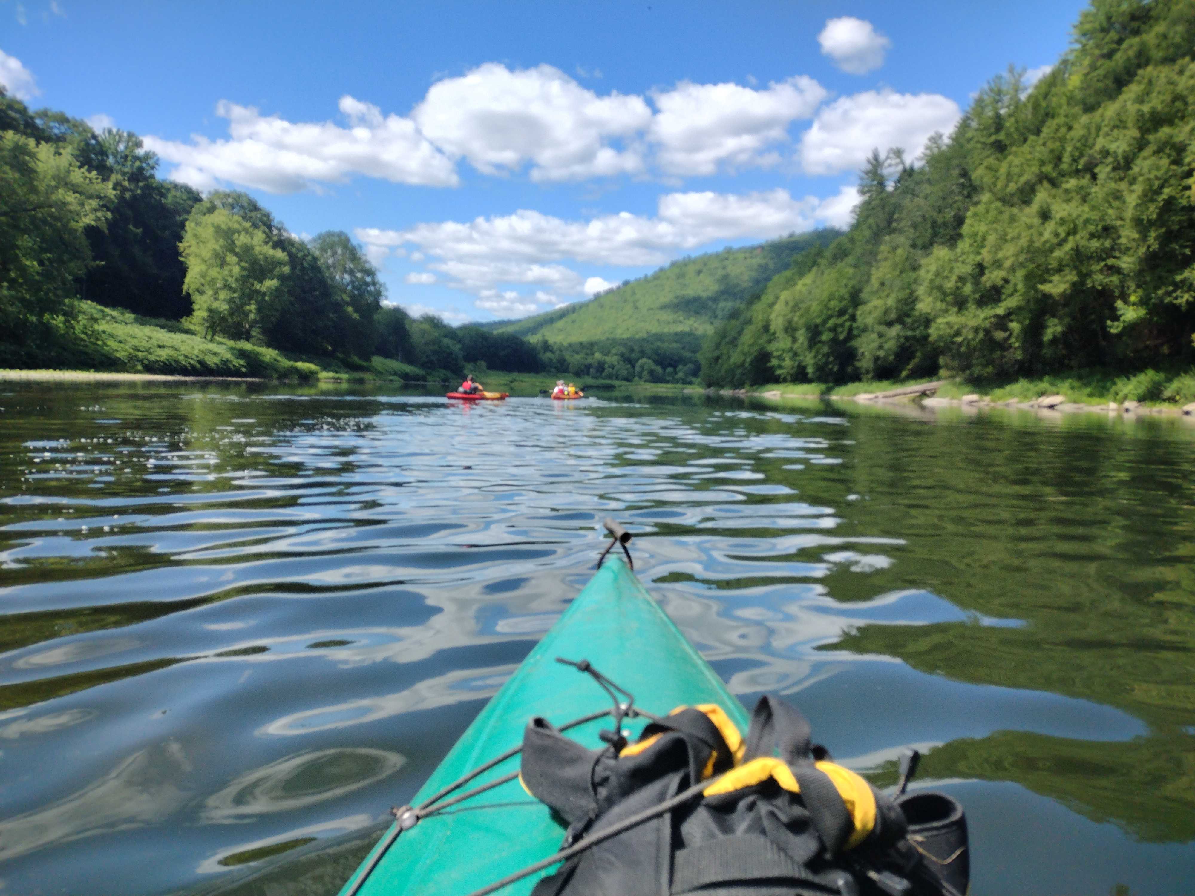 First person POV view on a blue-green kayak on a river. Riverbanks covered in trees.