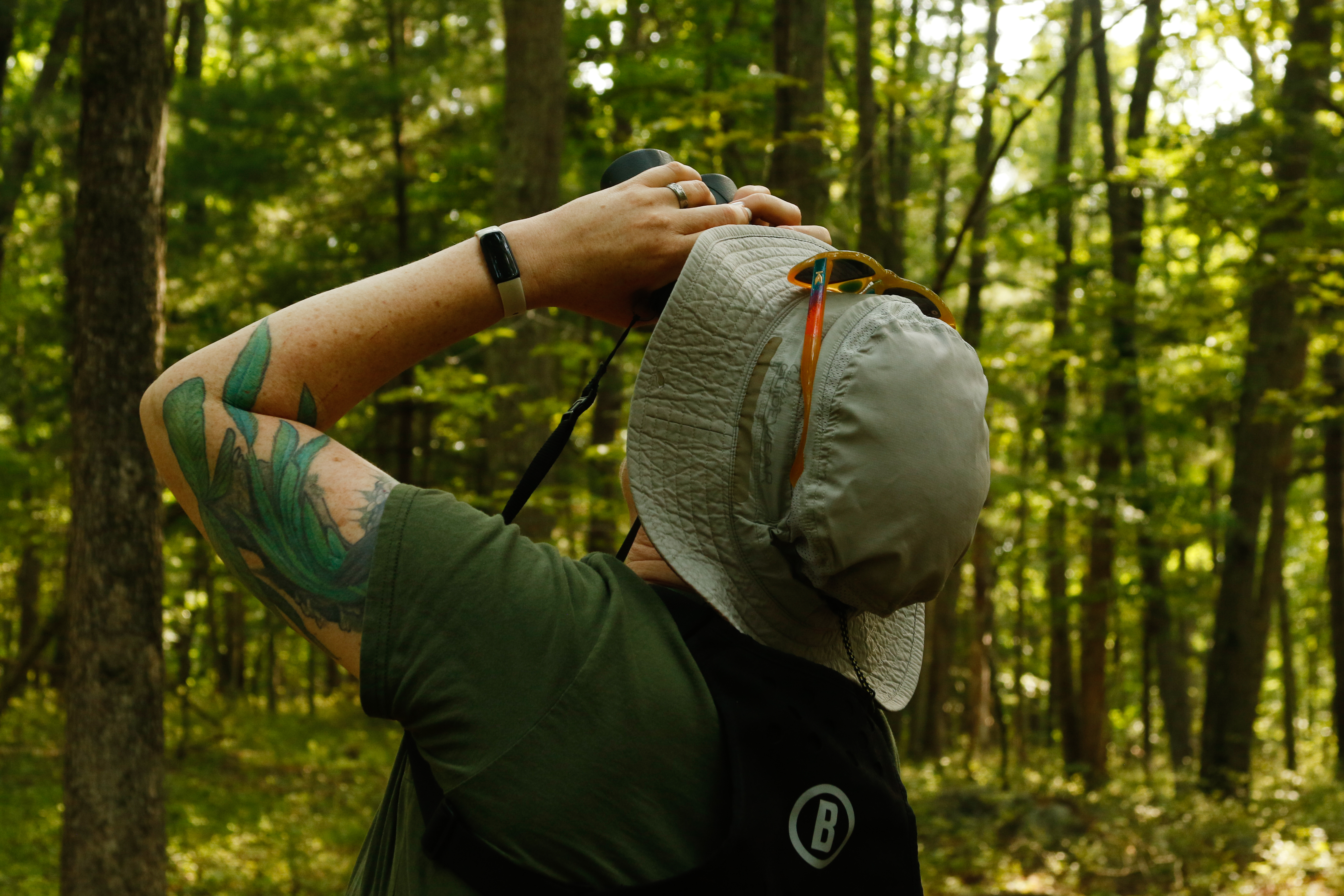Woman wearing bucket hat looks upwards with binoculars. She is in a forested area.