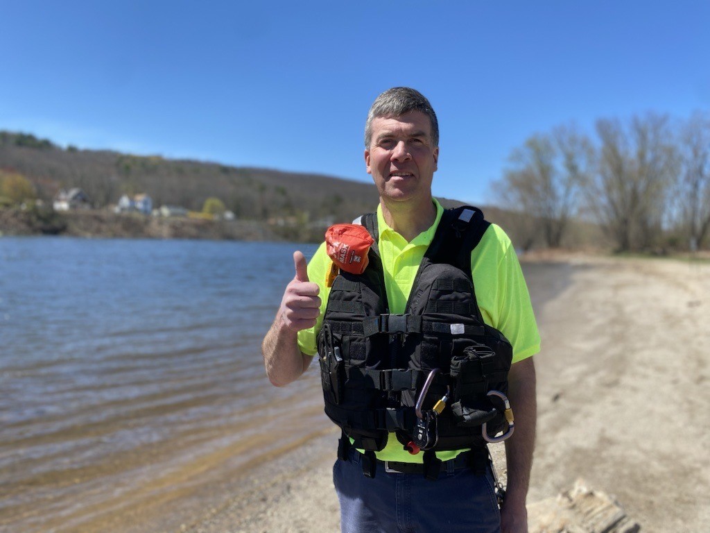 Man with salt and pepper hair smiles at camera, making thumbs up sign. WEars a black life jacket over a neon yellow shirt. River is behind him.