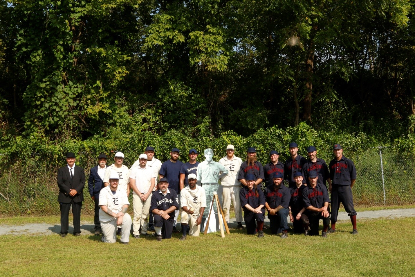 Two baseball teams of approximately 9 players each stand next to a cardboard standee of a young Zane Grey. One team wears a dark navy uniform with red lettering, the other with a mostly white uniform. In addition to the players are a couple referees.