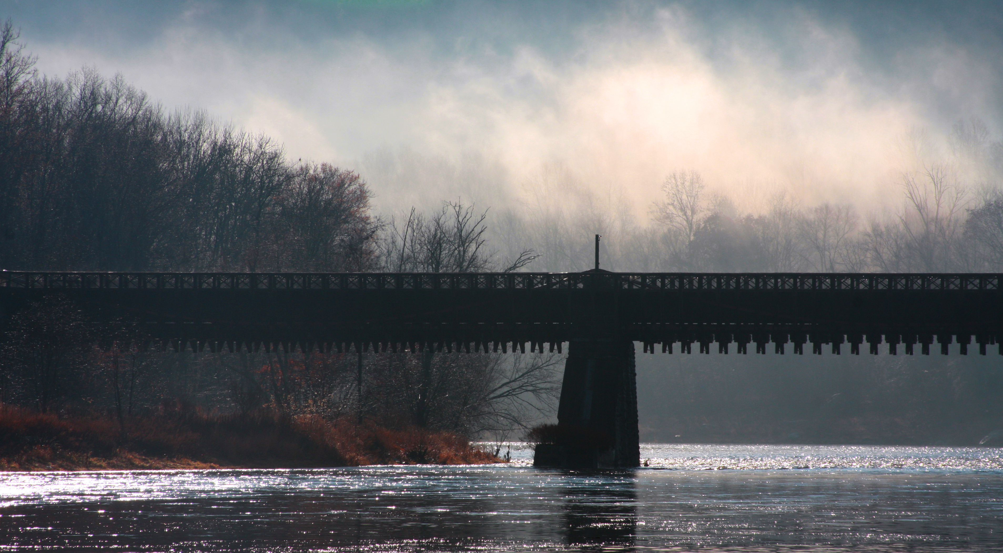 Section of a flat metal bridge over river, silhouetted against mist over the river. Mist is white in the sun against backlit hills. Light shines off river.