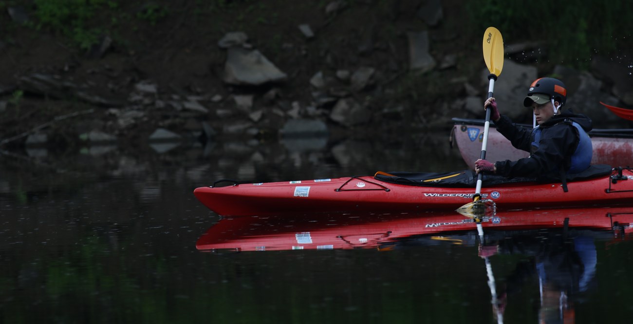 boy in helmet and life jacket cuts through water in bright red kayak.