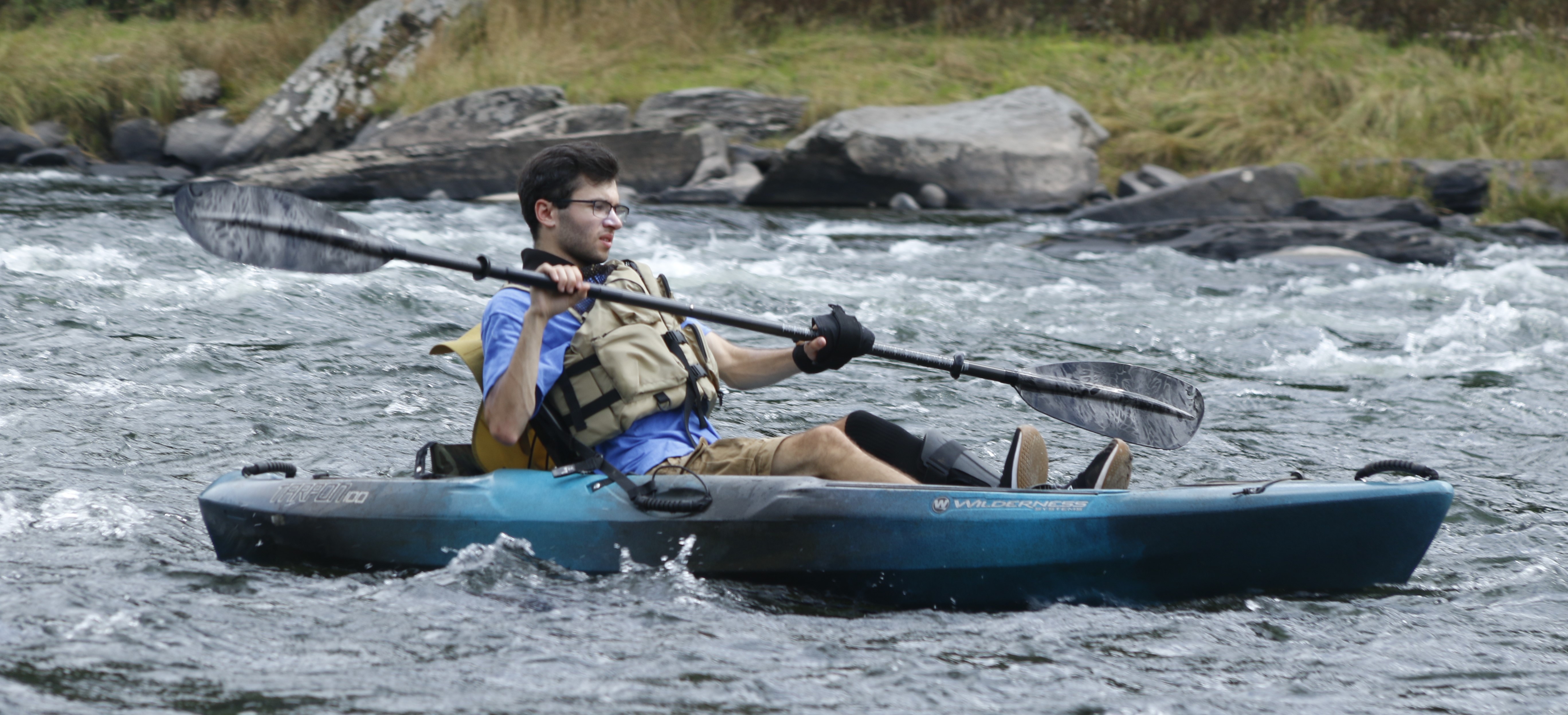 Young man paddling in sit-upon kayak in the river with white rapids. One of his legs is in a brace. He is wearing a life jacket.