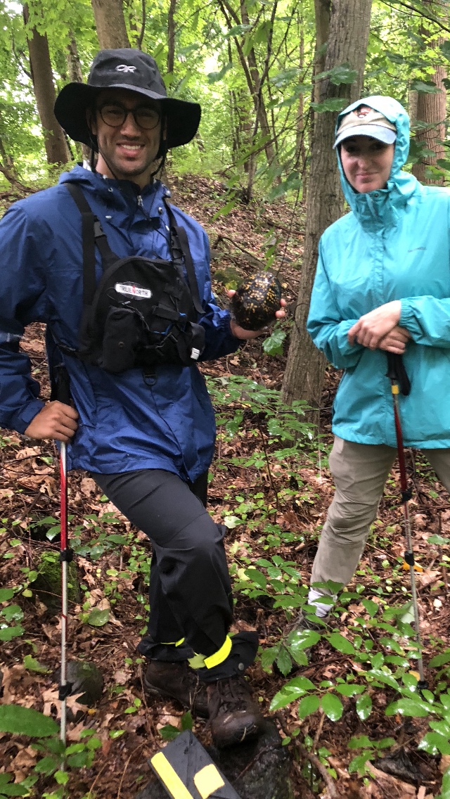 two interns stand smiling side by side in the forest. one is holding a turtle.