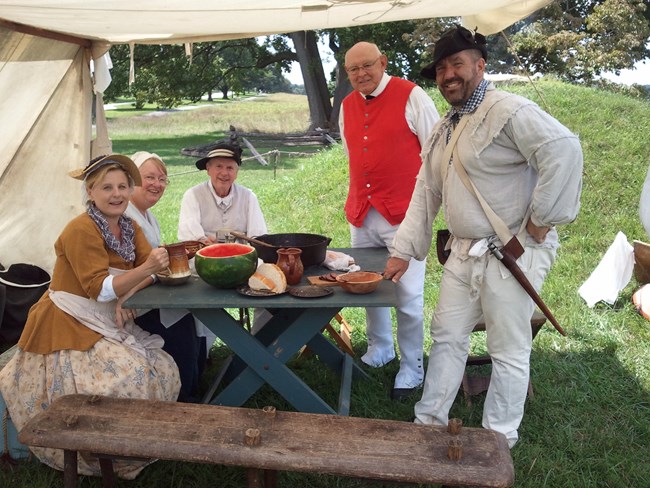 A group of people dressed in period attire stand under a lean-to. A group of people dressed in period attire stand under a lean-to.