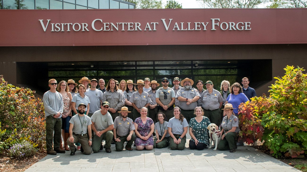 smiling men and women in wearing park ranger uniforms stand together in front of the Visitor Center at Valley Forge