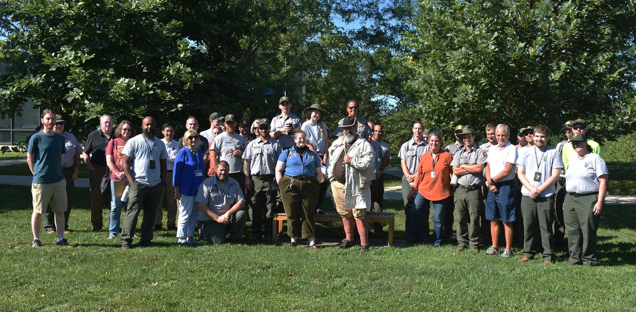 a group of smiling people, some wearing uniforms, stand on grass