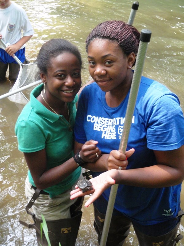 photograph, two YCC crew members stand in a shallow stream and hold a turtle