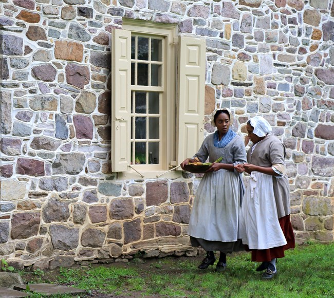 photograph, outdoors, stone wall, window, two ladies talking in eighteenth century clothing