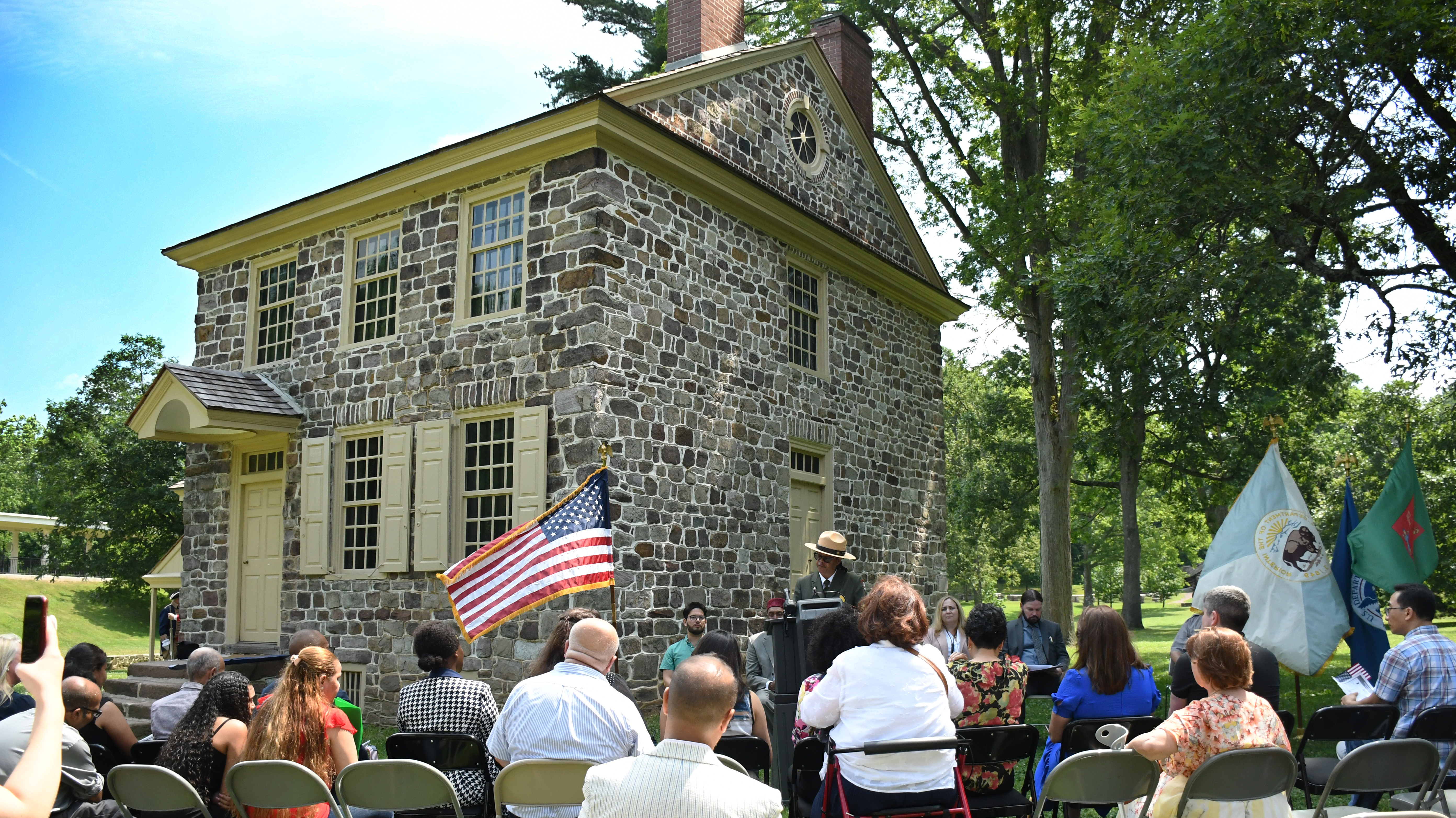 Valley Forge Superintendent Rosalyn Fennell Addresses New Citizens a park ranger at a podium next to a small stone farmhouse speaks to people seated in front