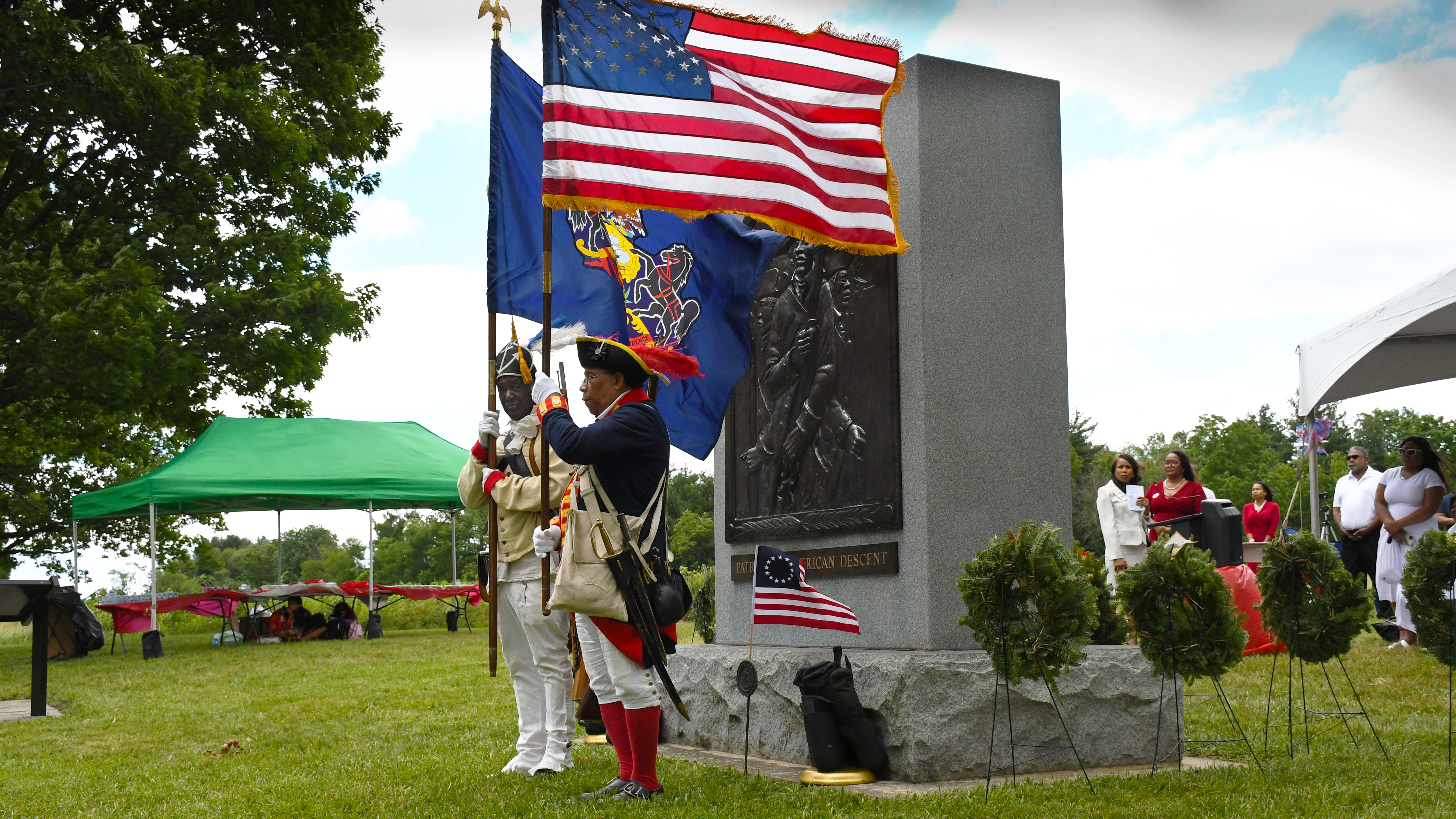 Two black soldiers in 18th century uniform stand next to the stone monument with flags
