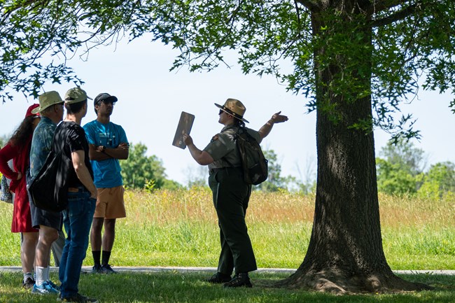 A uniformed park ranger speaks to a group under a tree