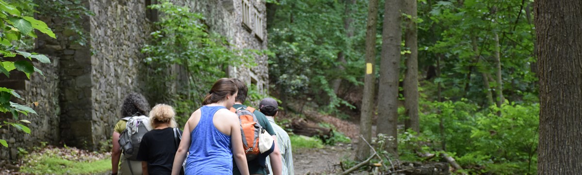 Hikers travel up hill on a wooded trail past stone ruins. Hikers travel up hill on a wooded trail past stone ruins.