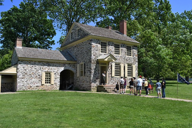 a small stone farmhouse. a park ranger stands at the front door and smiles at a group of people on the sidewalk in front of the house