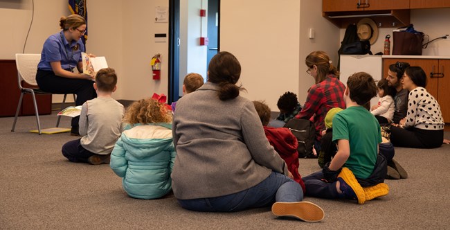 A woman seated in a chair holds a book up to a group of children and adults seated on the floor.