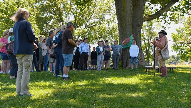 A ranger in 18th century clothing speaks to a crowd under a tree