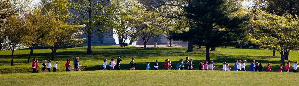 A line of runners pass across a grassy area in front of a concrete arch. A line of runners pass across a grassy area in front of a concrete arch.