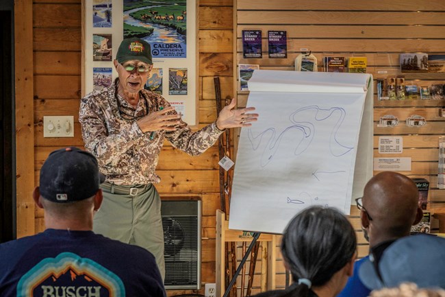 A man wearing a camouflage shirt addresses a seated audience inside a building.