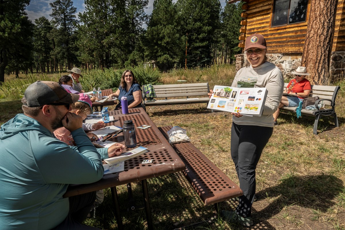 An artist holds up a sketchbook with watercolor paintings for a group of people at outdoor picnic tables.