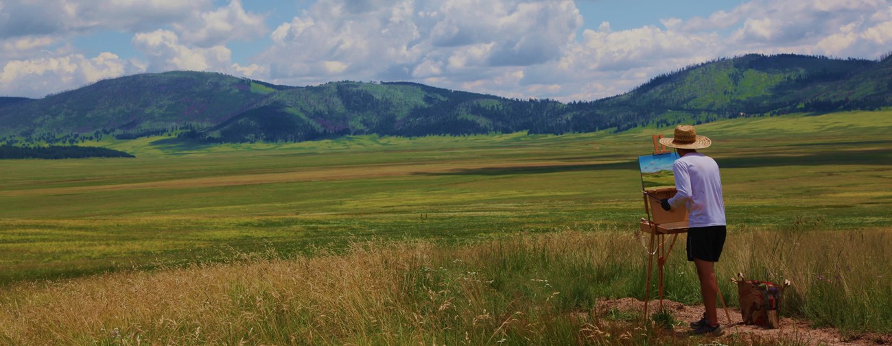 An artist wearing a wide-brim hat paints on a canvas on an easel in a vast, grassy valley.