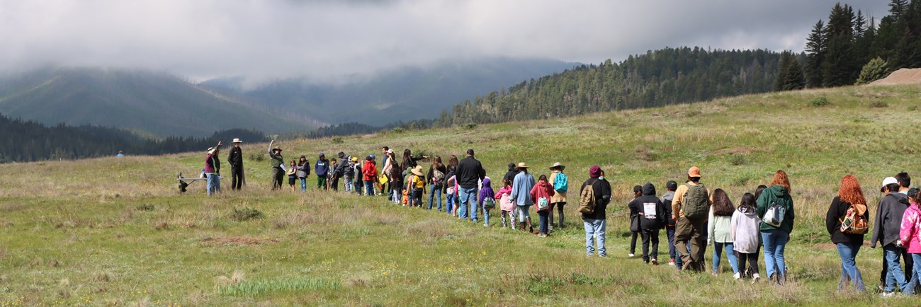 A long line of students and chaperones hike a trail single-file behind a ranger.
