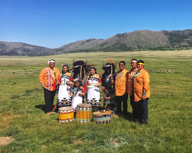 A group of indigenous musicians and dancers wearing traditional buffalo dance costumes stands in a grassy valley.