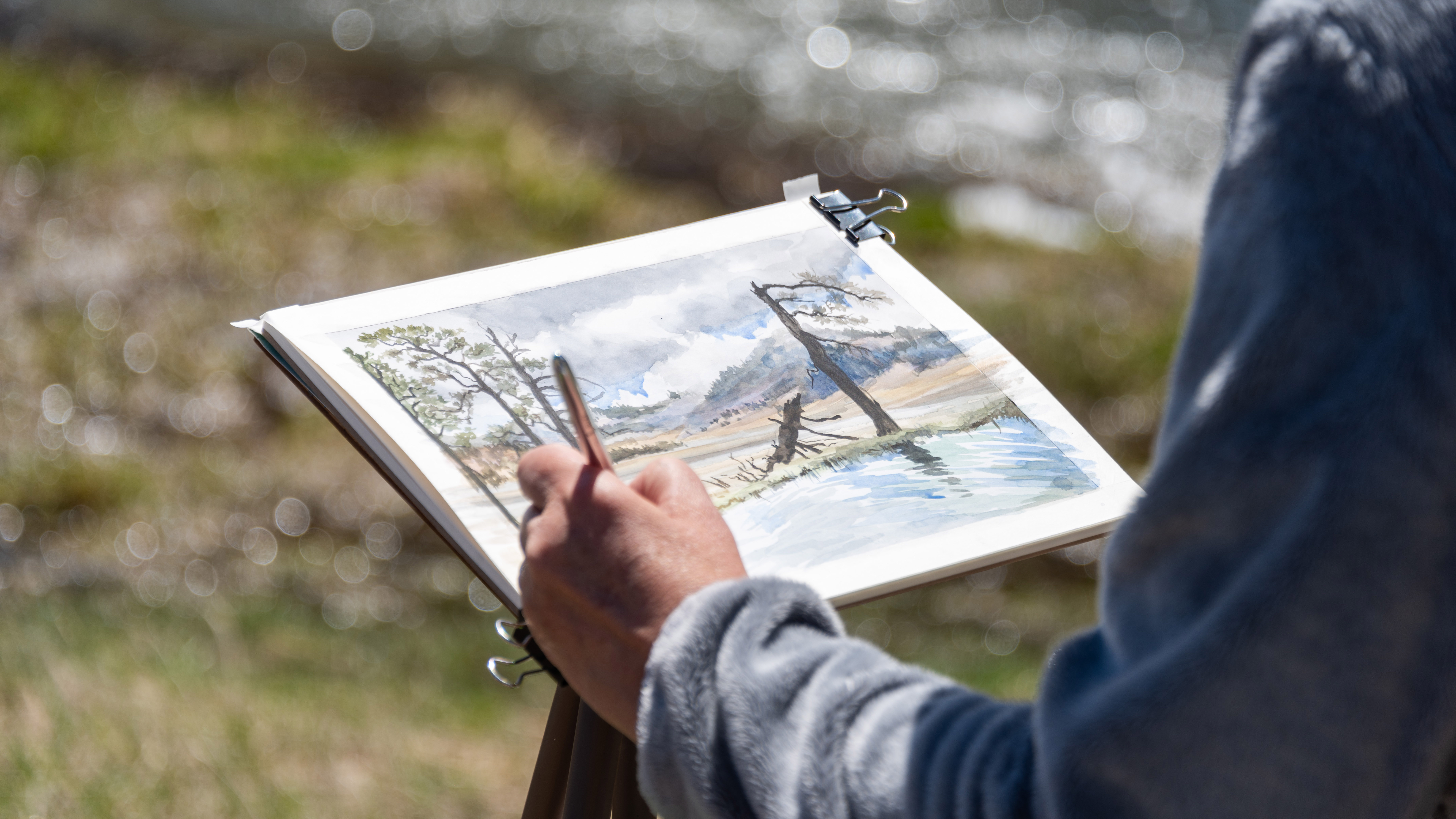 Close-up of an artist's left hand and arm as they paint a watercolor scene of a pond.