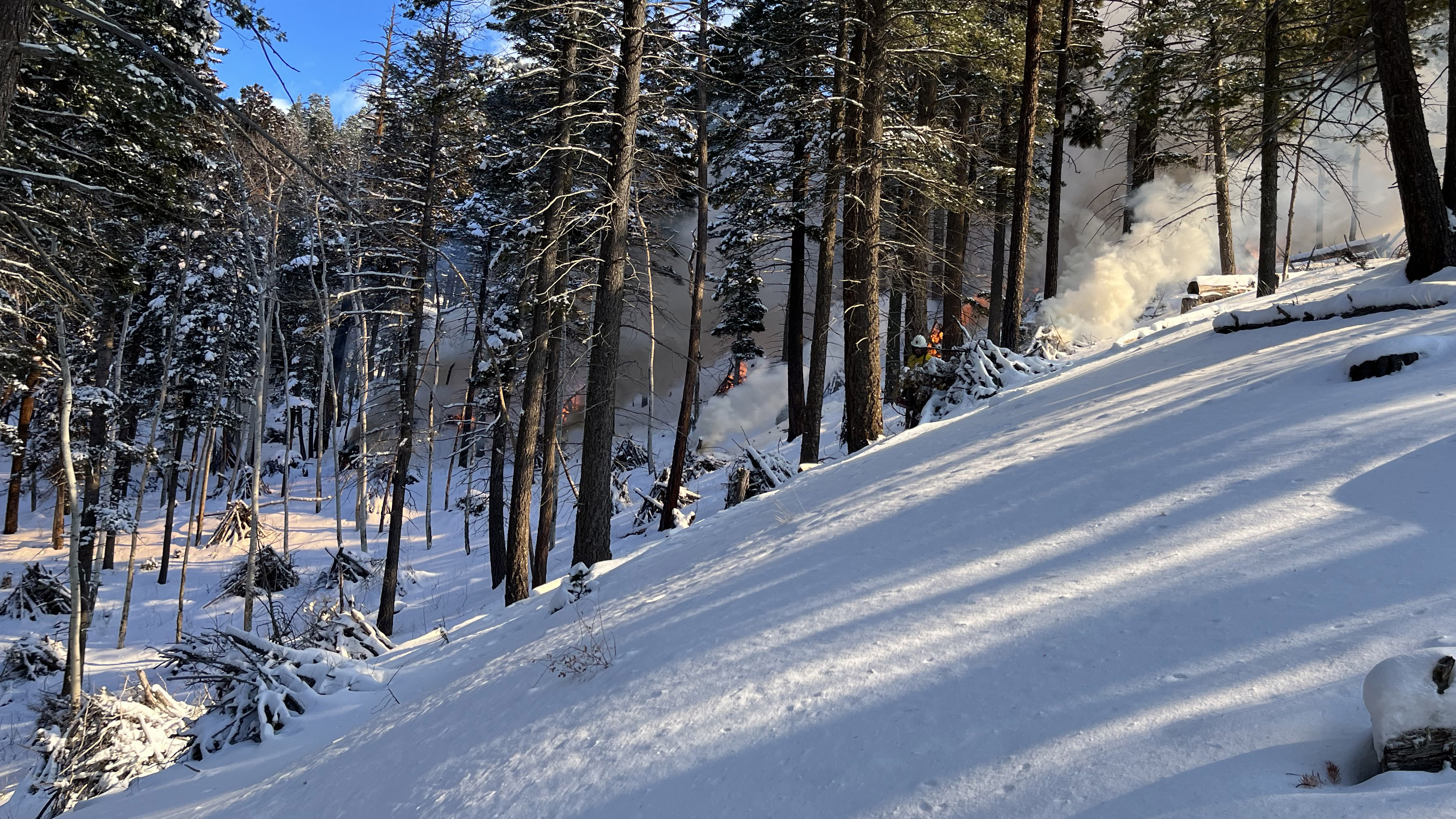 A fre crew member walks near burning piles on a snow-covered mountain slope.