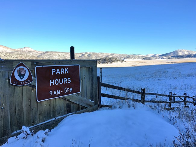 Valles Caldera National Preserve's entrance sign showing park hours 9 AM - 5 PM with 3 inches of snow in the background.