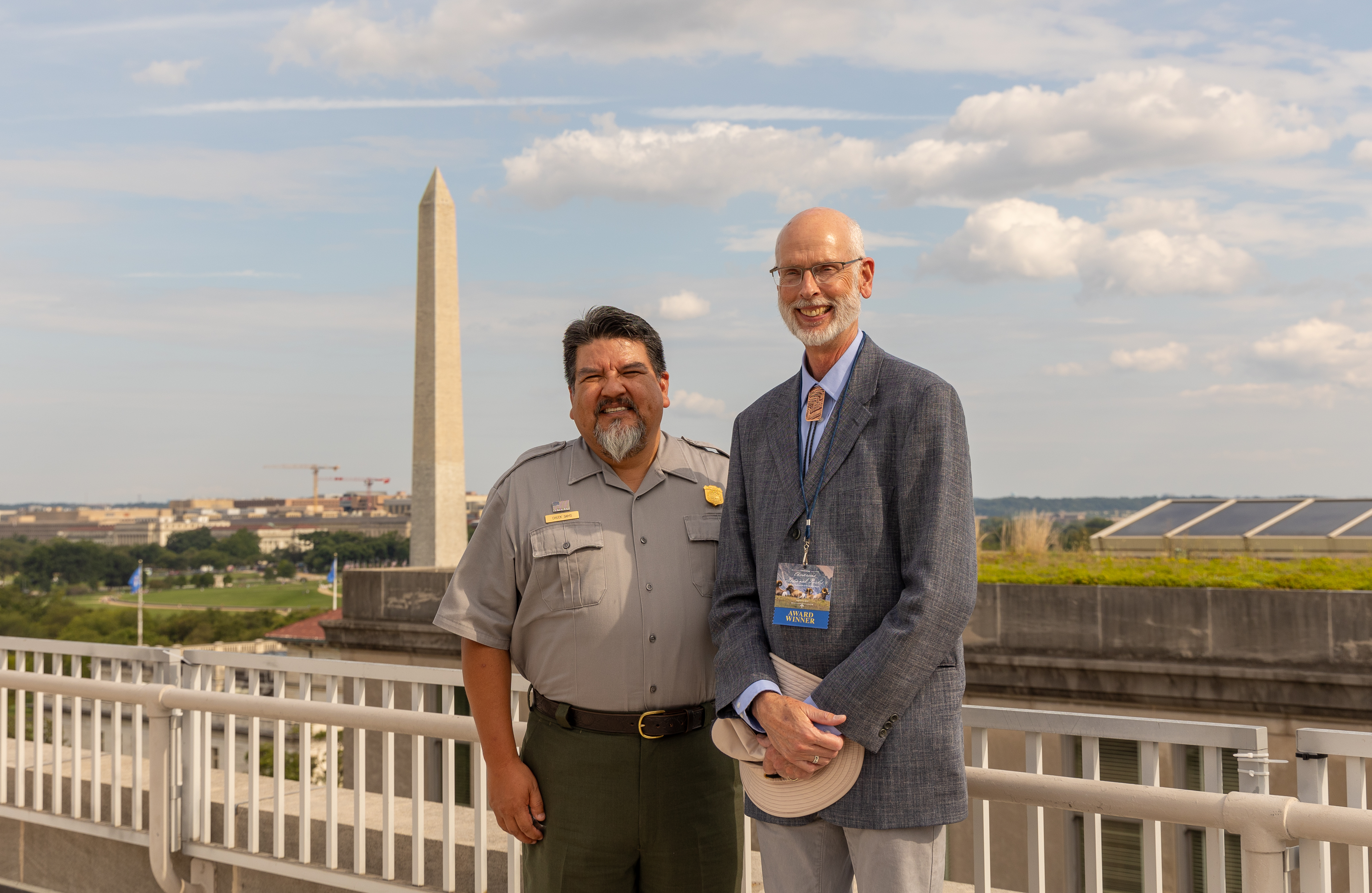 Two men stand on a rooftop balcony with the Washington Monument visible in the distance.