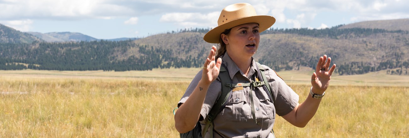 A park ranger gestures with both hands while addressing a group on a guided hike in a grassland.