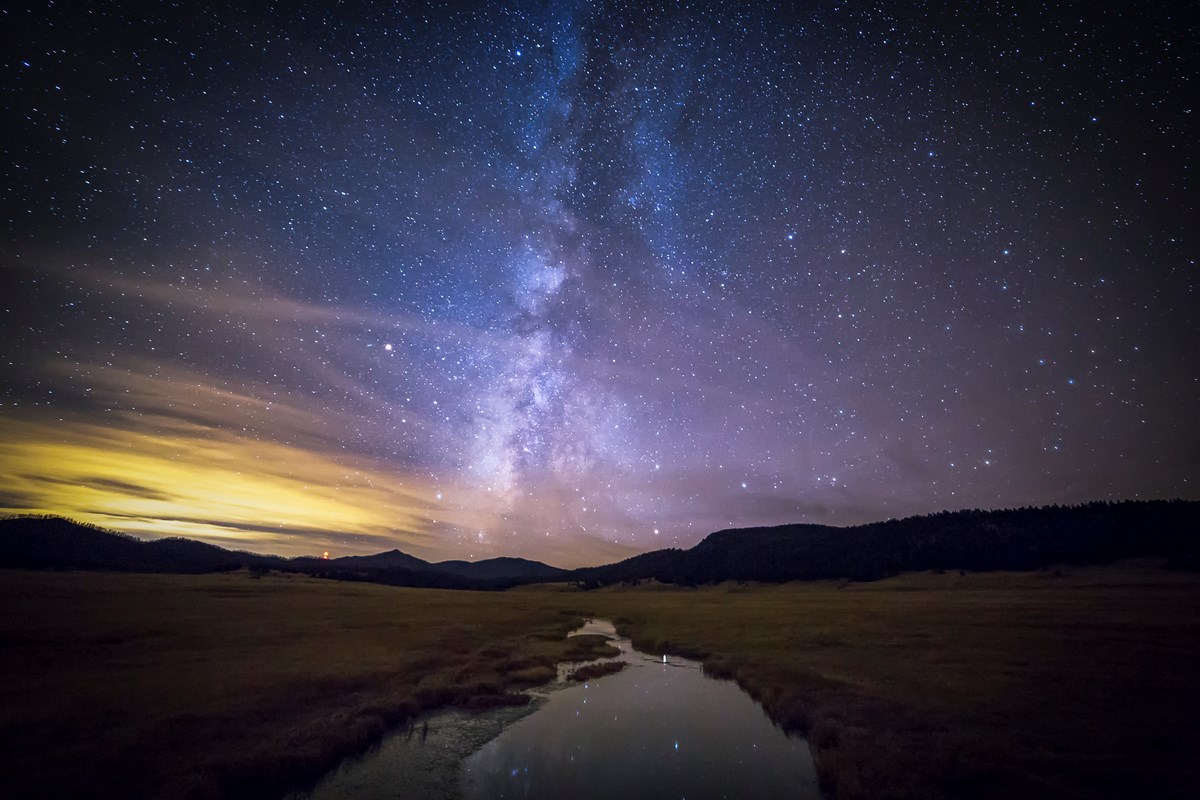 A creek runs through a wide grassy valley. It is nighttime with stars filling the clear sky. A silhouette of mountains defines the far away horizon. The night sky is reflected in the calm water.
