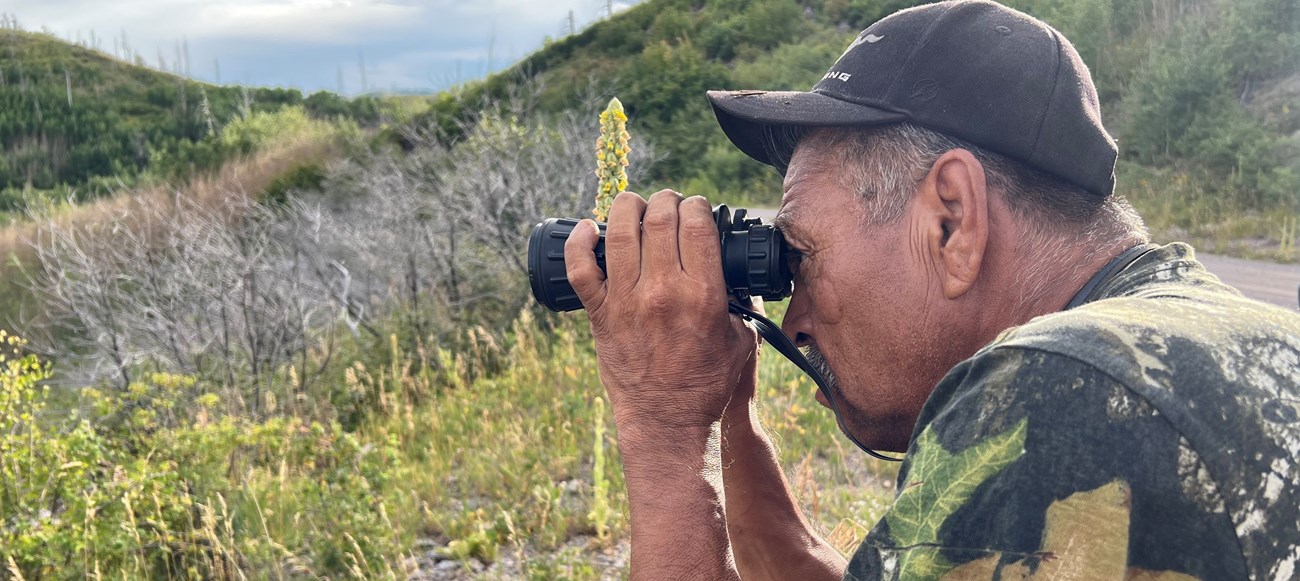 A man wearing camouflage uses binoculars to look across a mountainous landscape