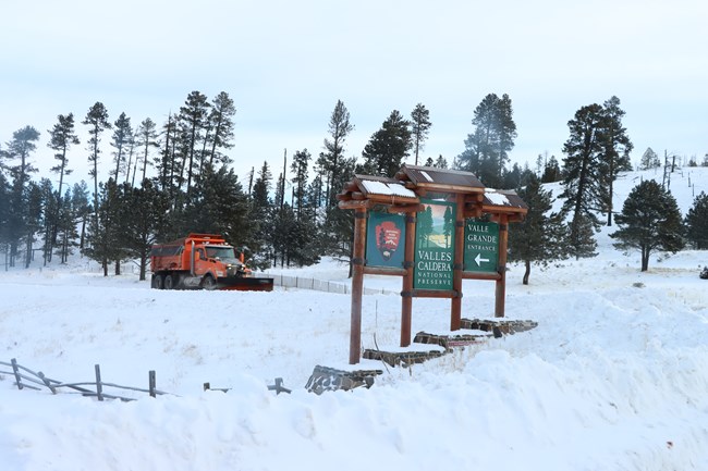 An orange snowplow truck drives past a snowy Valles Caldera National Preserve entrance sign.
