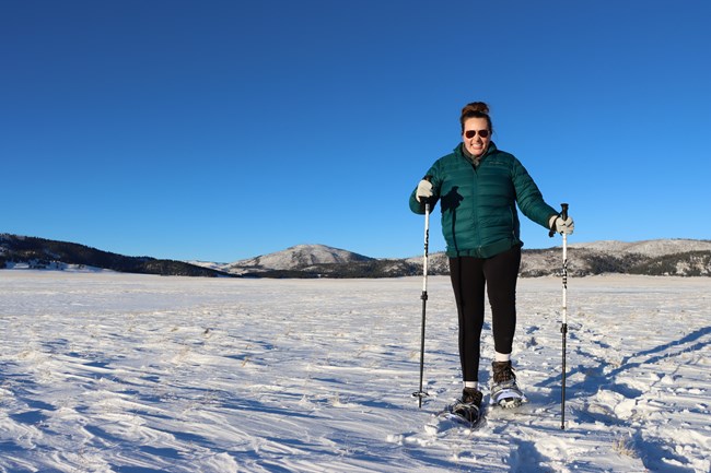 A woman wearing sunglasses and a green coat snowshoes toward us in a snowy valley.