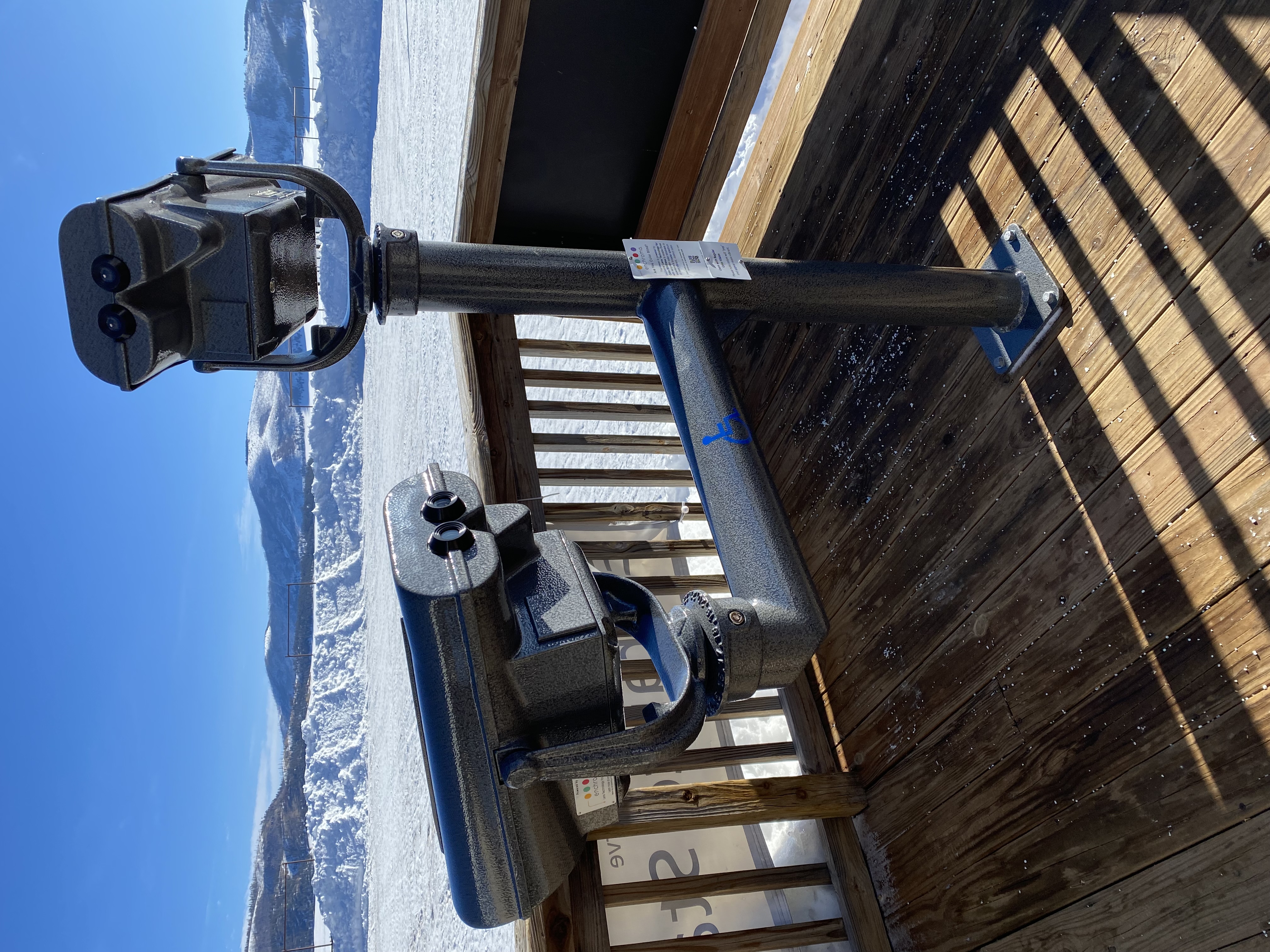 Two spotting scopes, one of which is labeled wheelchair accessible, are fastened into a wooden porch in a snowy landscape.