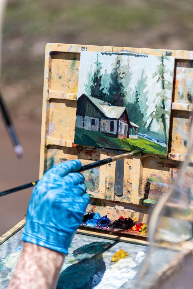 A gloved hand paints a picture of a log cabin.