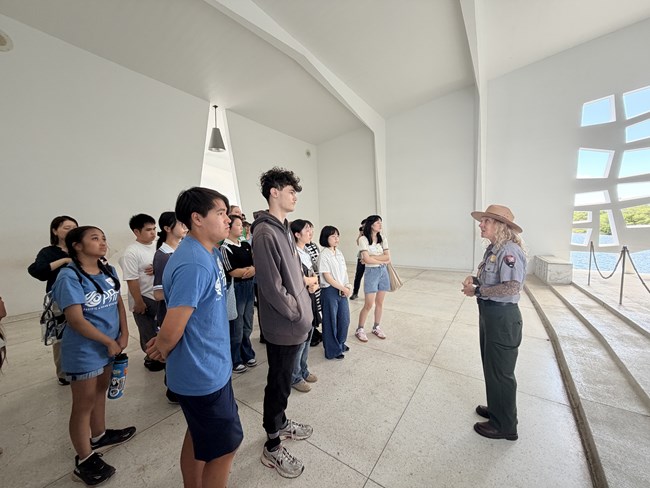 A ranger in a gray shirt, green pants, and brown flat brimmed hat stands in front of many students on a white floor at USS Arizona Memorial