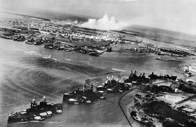 A black and white aerial photo of the line up of battleships, called Battleship Row, at the start of the Pearl Harbor attack.