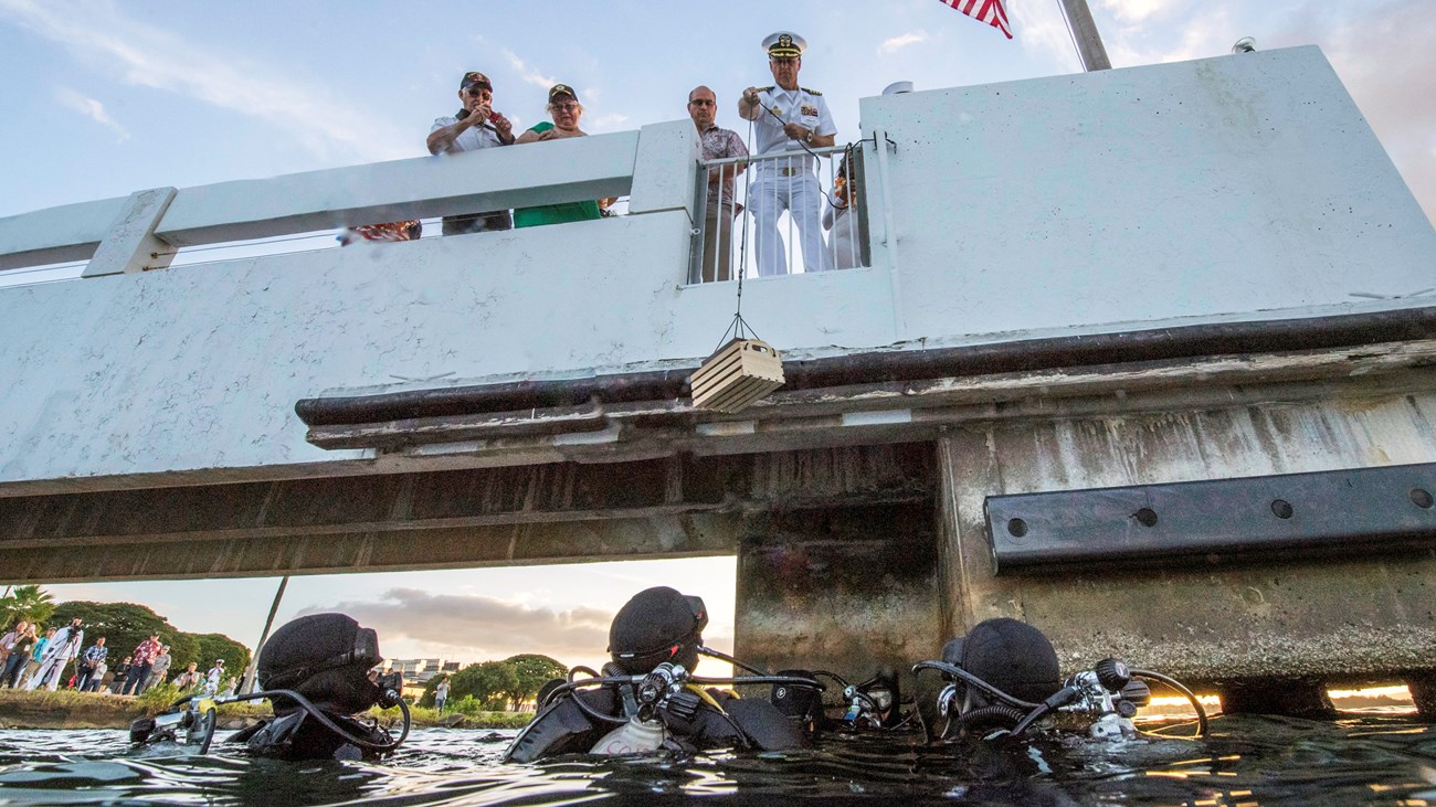 Three National Park Service divers in the water reach up for the urn of USS Utah crewmember Harry Hohstadt during his interment ceremony on December 6, 2017.