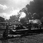 Sailors at Ford Island Naval Air Station look on as the USS Shaw explodes in the distance. This view is of the PBY ramp with assorted aircraft scattered among the debris.  Barely seen in the background is the beached USS Nevada.