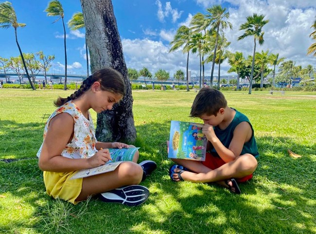 Two kids sit on the grass filling out booklets