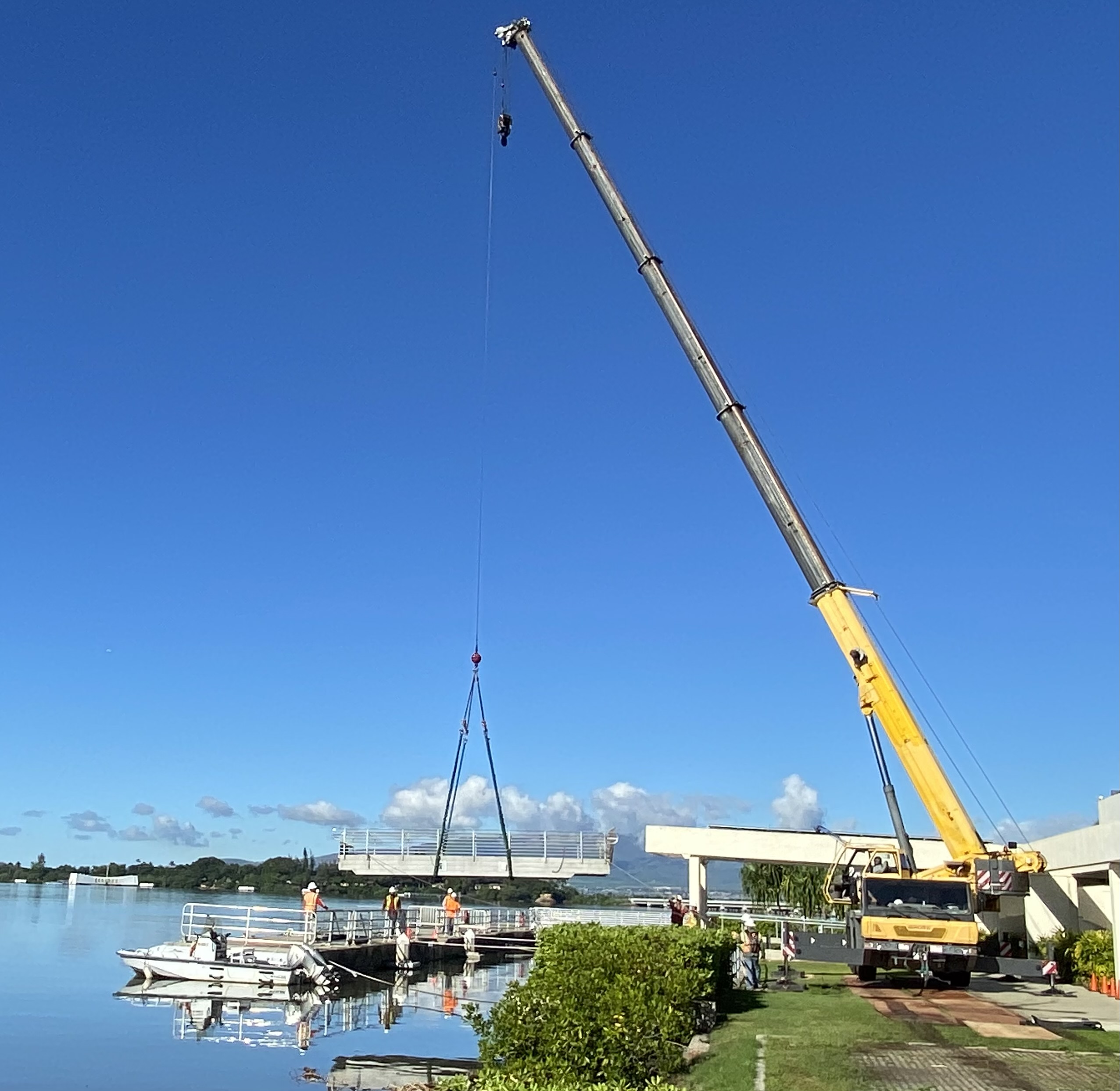 NAVFAC workers in hardhats and safety vests watch a crane lift the shoreside ramp into place.