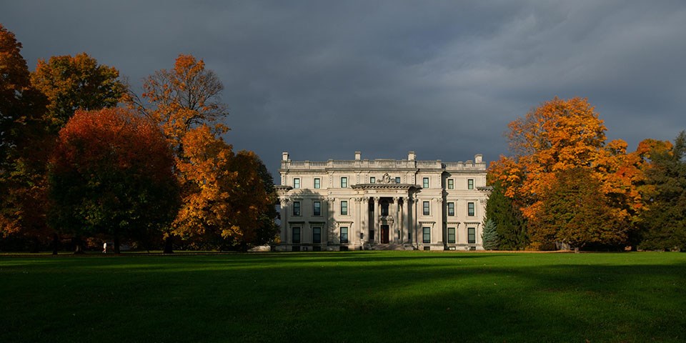A grand five story house amid trees with orange leaves on a green lawn.