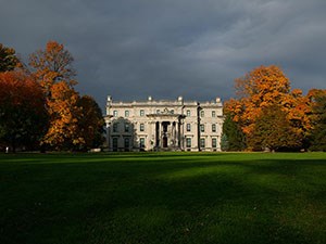 A grand five story house amid trees with orange leaves on a green lawn.