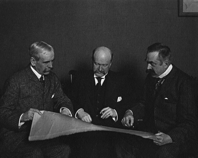 Three men in suits looking over a large document.