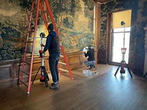Two conservators carefully remove a tapestry from a wall.