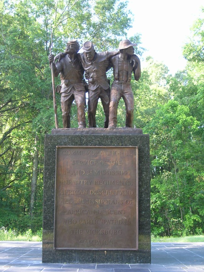 Statue of three African American Civil War soldiers. Two on either side, supporting a third in the center. One carries a rifle. Statue is on a dark colored base with a metal plaque. Green trees in background